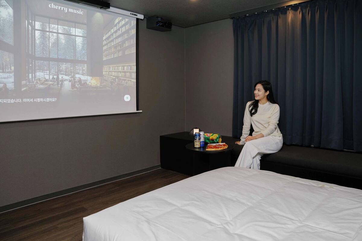 Habitación de hotel con cama, una mesita con aperitivos, pantalla de proyector mostrando una presentación y un ventanal con cortinas.