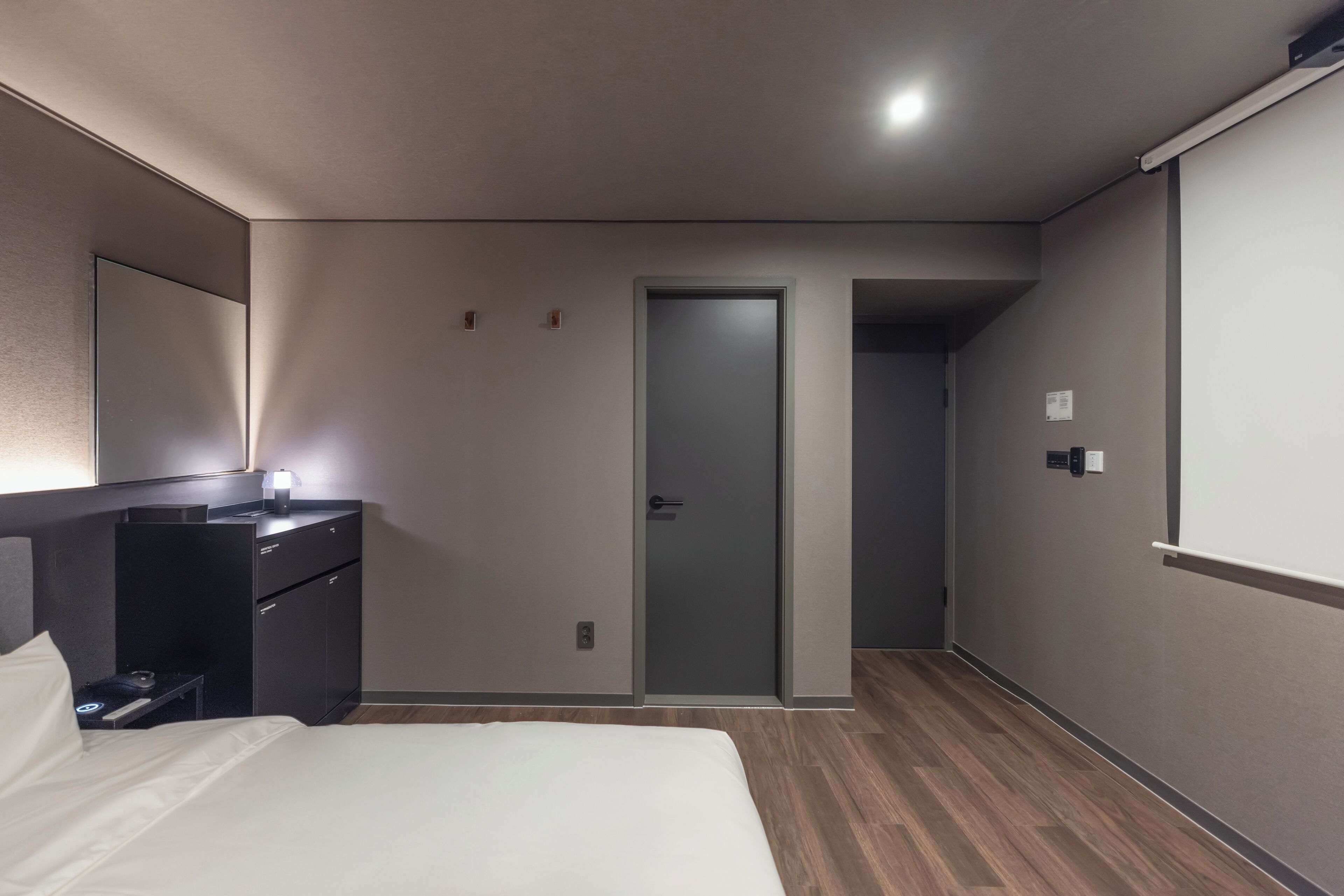 A modern hotel room with a bed, a bedside table, a mirror, a wardrobe, and a door leading to another room.