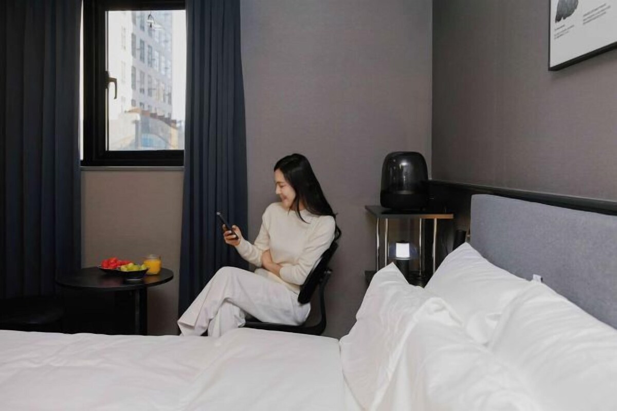Een hotelkamer met een bed, een stoel, een klein tafeltje met fruit en een fles, en een raam met gordijnen.