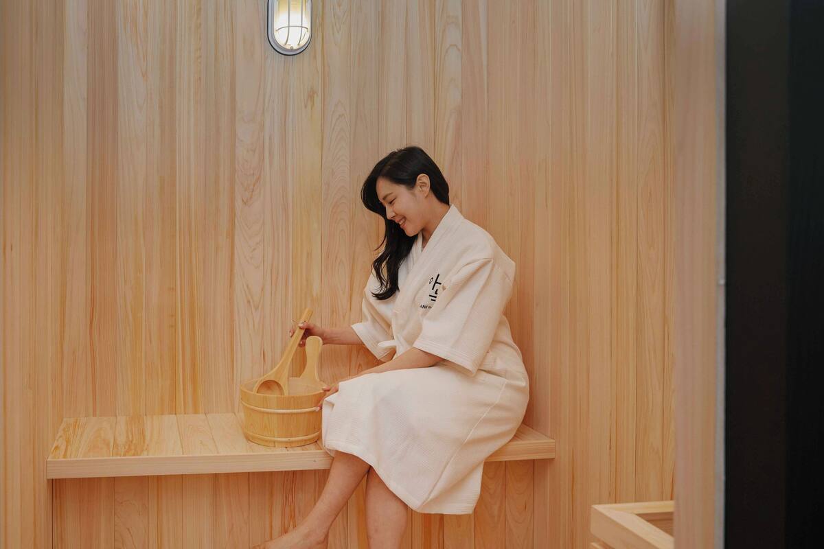 Una persona sentada en una sauna de madera, con un balde de madera y un banco.