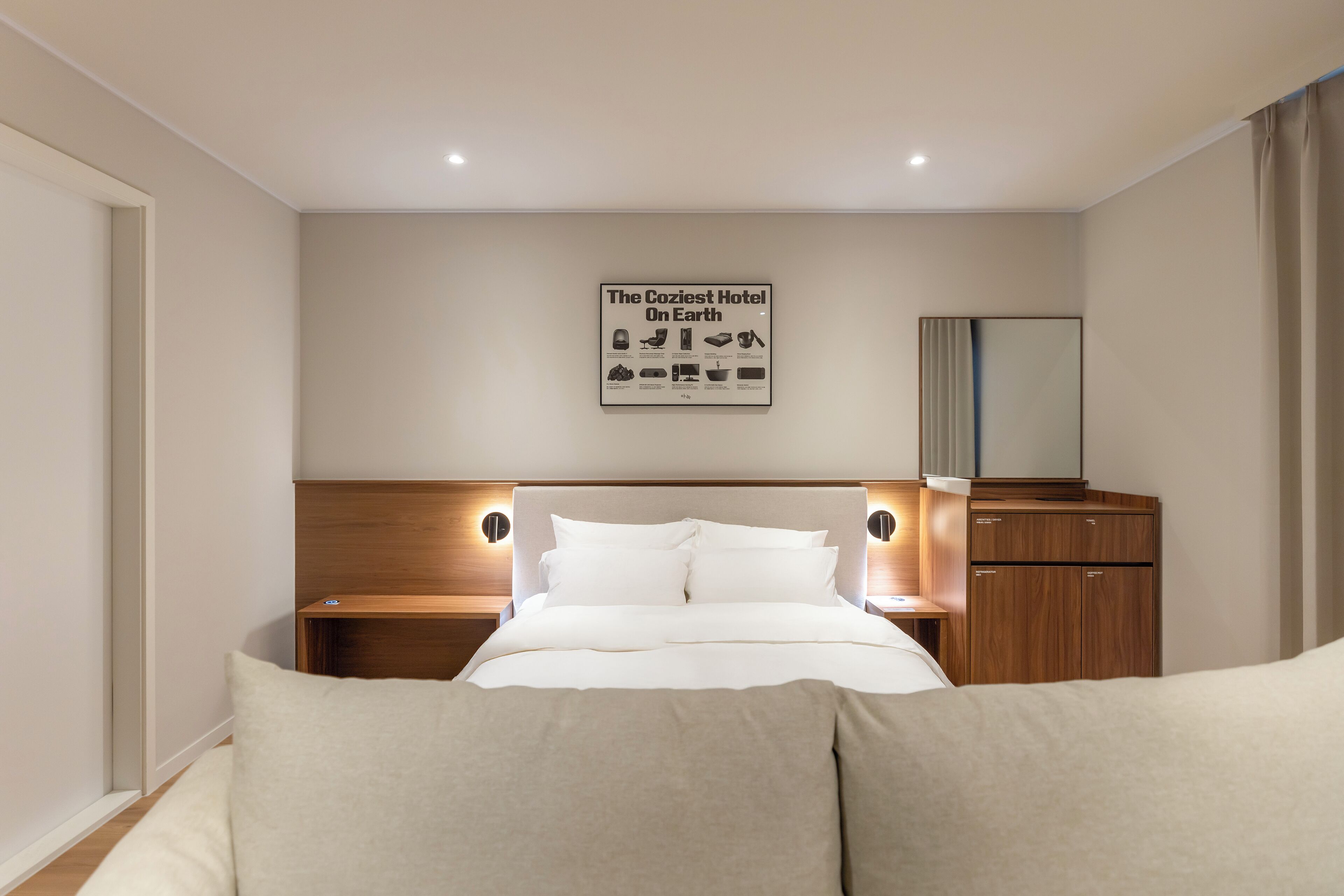 A modern hotel room with a large bed, wooden headboard, bedside tables, a nightstand, a mirror, and a framed picture on the wall.