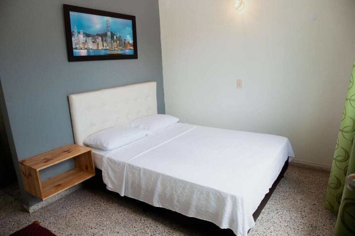 Een enkel bed met een wit hoofdeinde en beddengoed, een houten nachtkastje, een ingelijst beeld aan de muur en een groen gordijn.