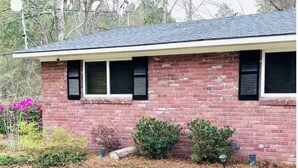 Een gelijkvloers bakstenen huis met plat dak, een veranda en een kleine voortuin met struiken en bloembedden.