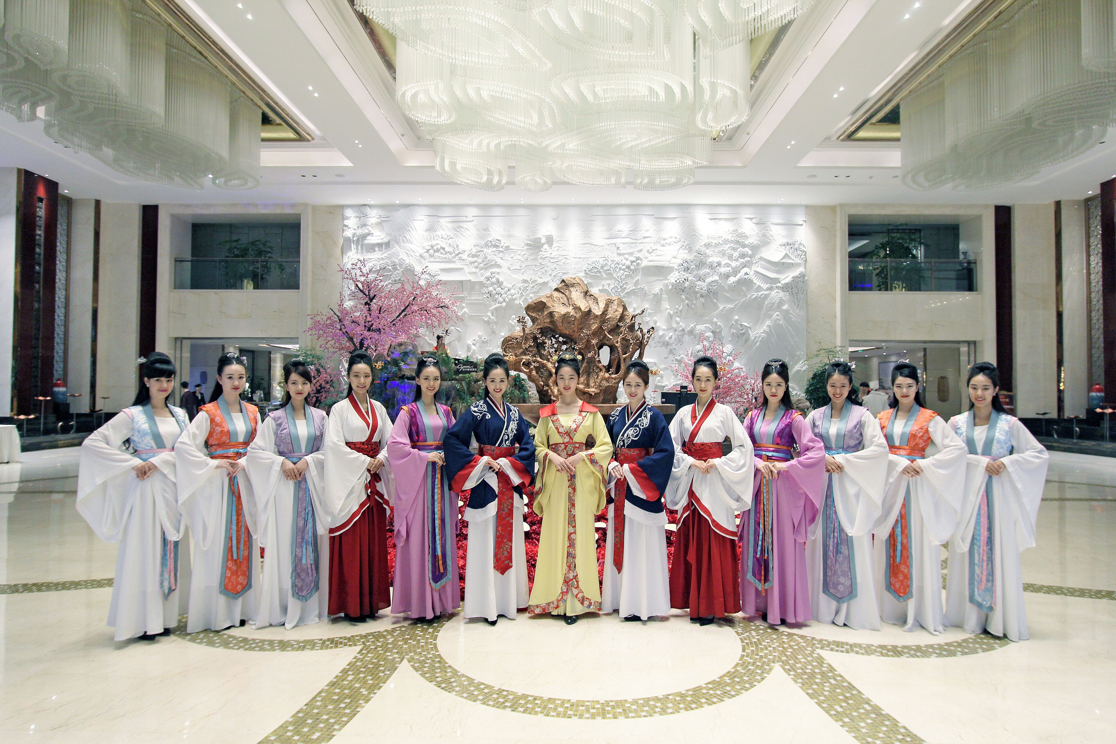 A group of people in traditional attire standing in a spacious, well-lit lobby with a decorative backdrop and a large chandelier.