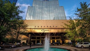 A hotel with a fountain in front, surrounded by trees and parked cars.