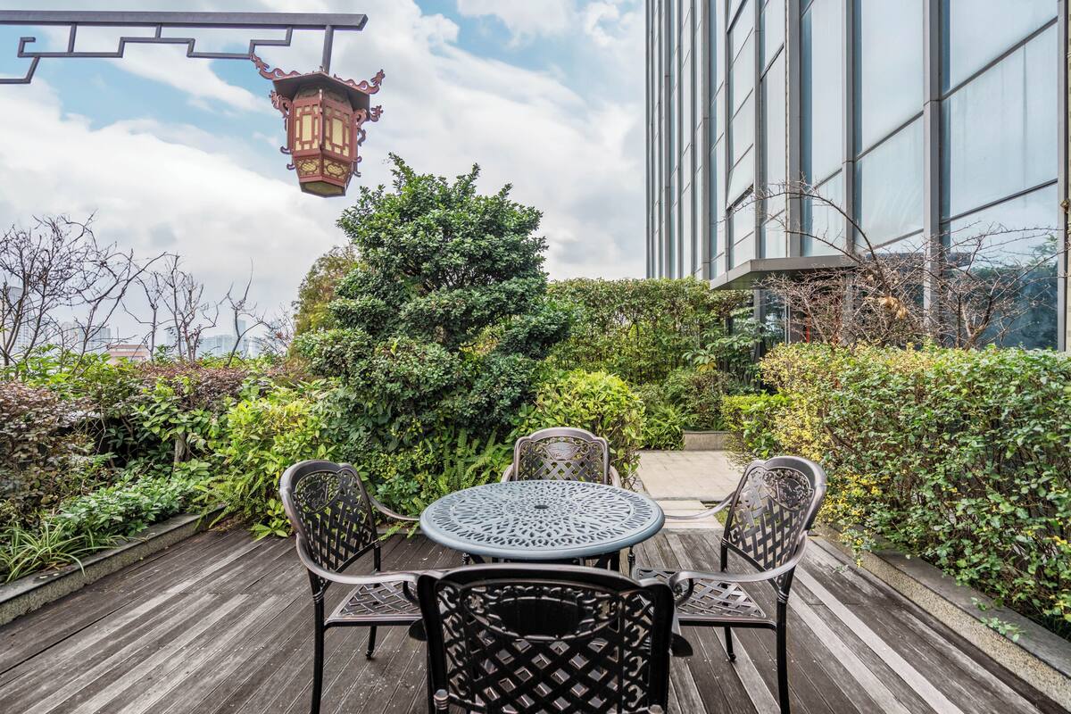 A wooden deck with a round table and chairs, surrounded by greenery and a building with large windows.
