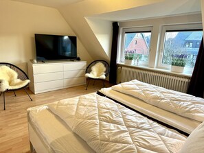 A bedroom with a TV on a dresser, two chairs, a bed with white bedding, and a window with a view of buildings and trees. - Haus Moin - Family-friendly vacation home for 7 people with terrace, car (Wenningstedt-Braderup (Sylt))