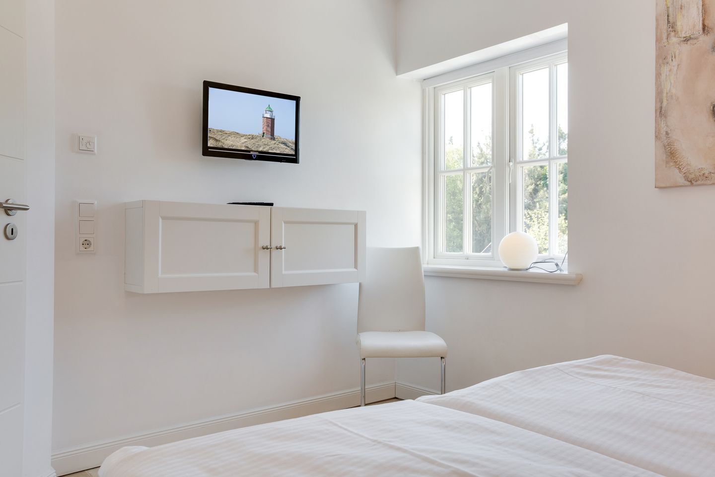Een slaapkamer met een bed, een witte stoel, een aan de muur bevestigde televisie en een raam met uitzicht op bomen.