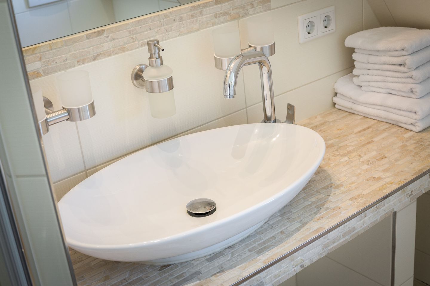 A bathroom with a white sink, chrome faucet, and soap dispenser. There are stacked white towels on the counter.