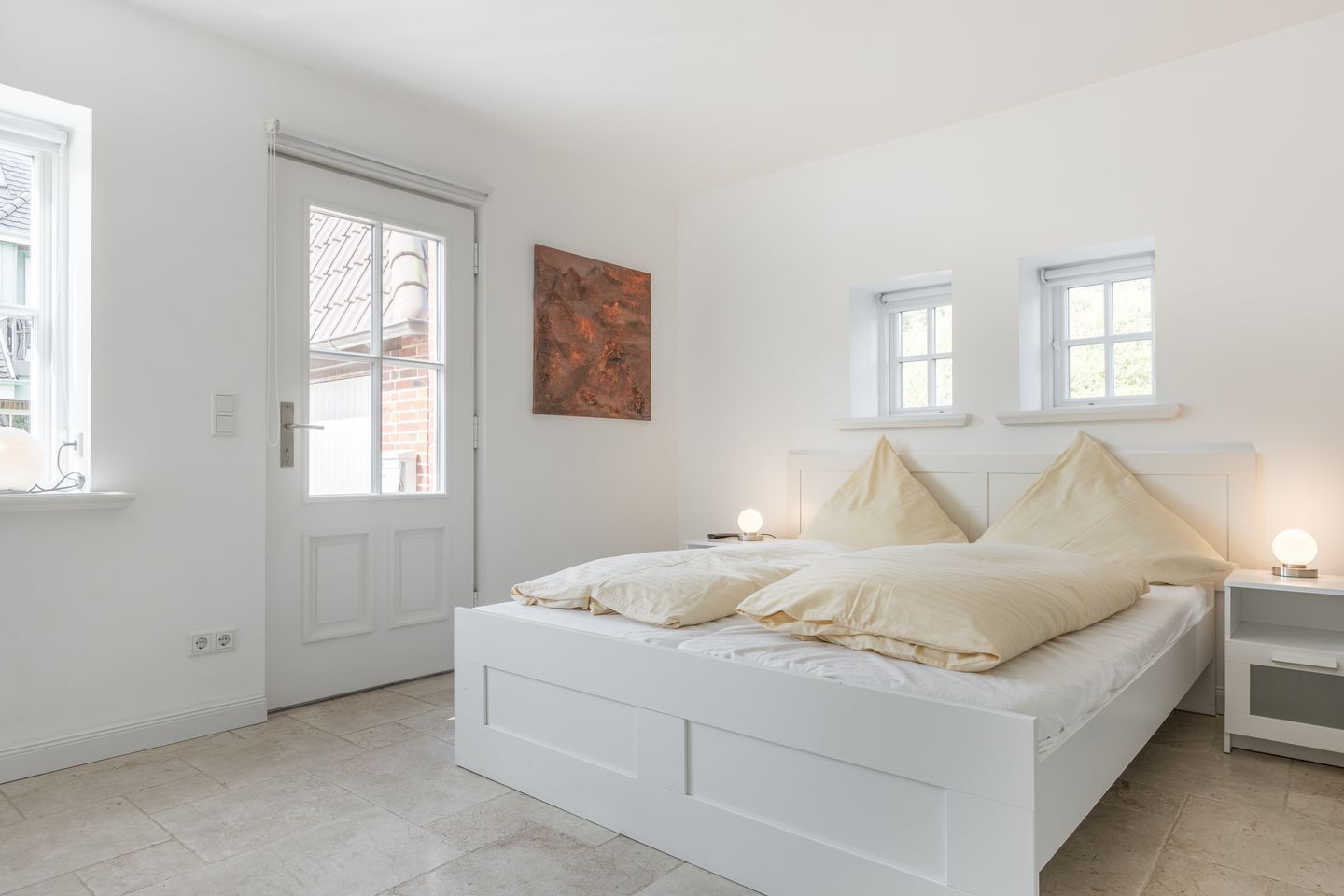 Une chambre à coucher avec un lit blanc, deux tables de chevet avec des lampes, une porte et deux fenêtres.
