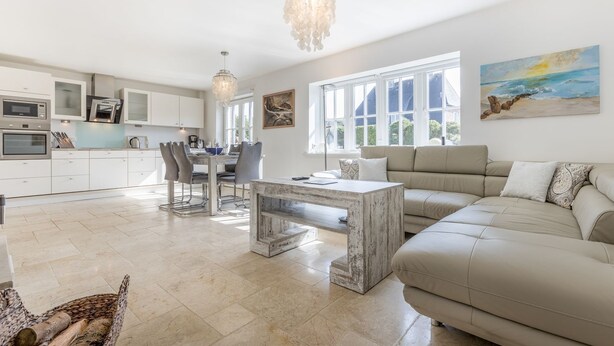 Eine moderne Küche mit weißen Schränken, ein Essbereich mit Tisch und Stühlen, ein beiges Sofa und ein großes Fenster mit Blick ins Freie.