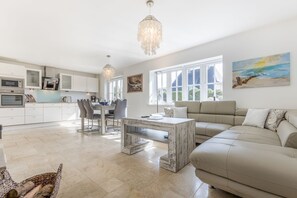 Een moderne keuken met witte kastjes, een eethoek met tafel en stoelen, een beige bank en een groot raam met uitzicht op het landschap.