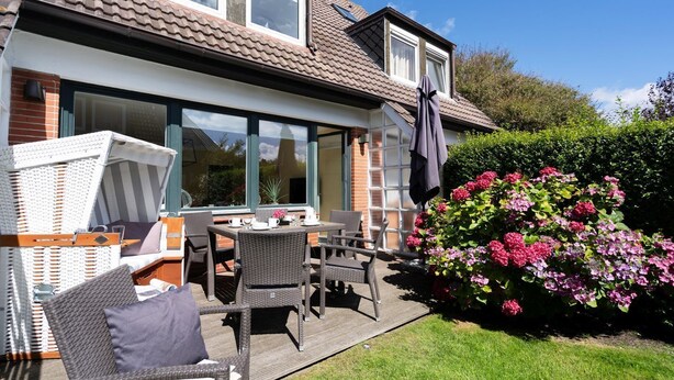 A patio with wicker furniture and a table set for dining, a brick house with large windows, and a garden with pink flowers. - Meeresstern - Beautiful house for 6 pers. with terrace, parking space, WLAN. (Westerland)