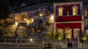 A building with a red facade and arched windows, situated next to a stone wall and staircase leading up a hill. - Villa with heated pool located in a unique position (Modica)