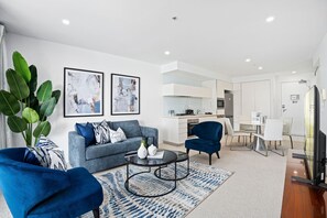 Un salon moderne avec un canapé gris, des fauteuils bleus et une table basse. Deux tableaux abstraits ornent le mur, ainsi qu'une grande plante verte. On aperçoit également un coin cuisine à l'arrière-plan.