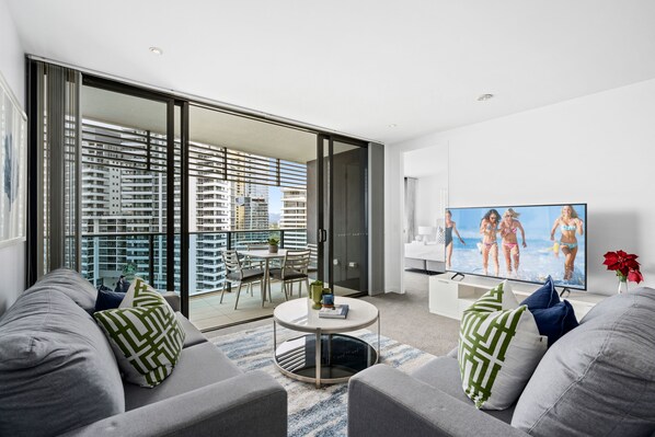 A modern living room with a large TV, grey sofas, a coffee table, and a balcony with a city view.
