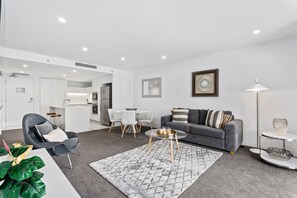 A modern living room with a grey sofa, a dining table with chairs, a kitchen area, and a large window.