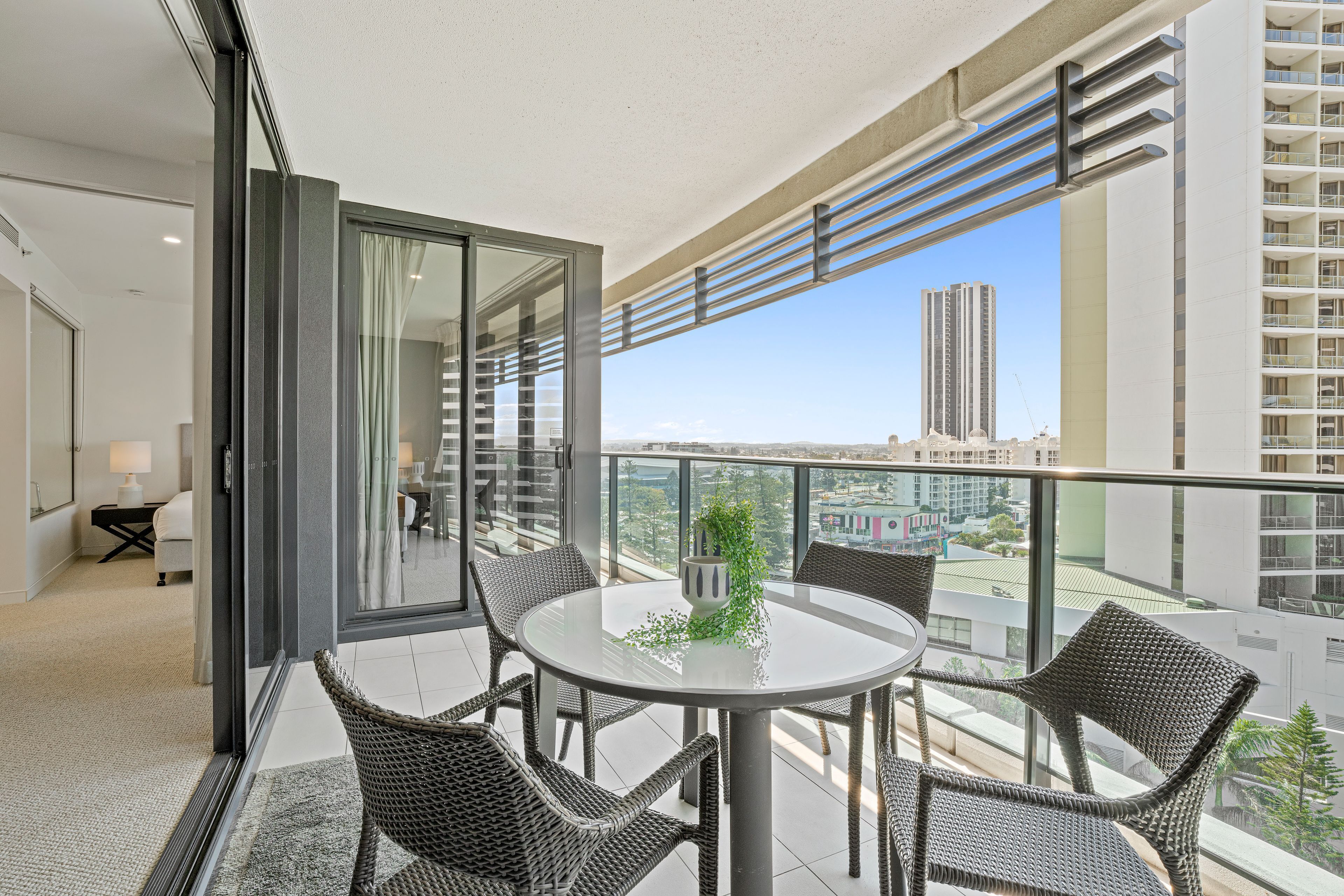 Un balcón con una mesa redonda y sillas de mimbre, con vista a un paisaje urbano de edificios altos.