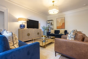 A modern living room with a blue sofa, a brown armchair, a glass coffee table, a white cabinet, a television, and a decorative shelf.