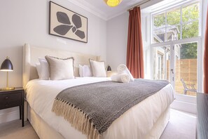 Una cama bien hecha con cabecera blanca, una lámpara de noche y una obra de arte enmarcada en la pared.