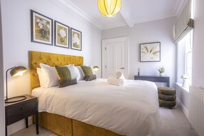 Un dormitorio con una cama grande, una mesita de noche, cortinas en la ventana e impresiones botánicas enmarcadas en la pared.