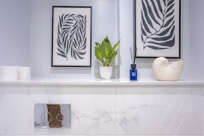 Un baño moderno con encimera de mármol, una obra de arte con hojas, una planta en maceta, una botella azul y una concha decorativa.