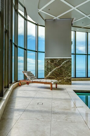 A sun lounger by a pool with a view of the sea, a marble wall, and large windows.