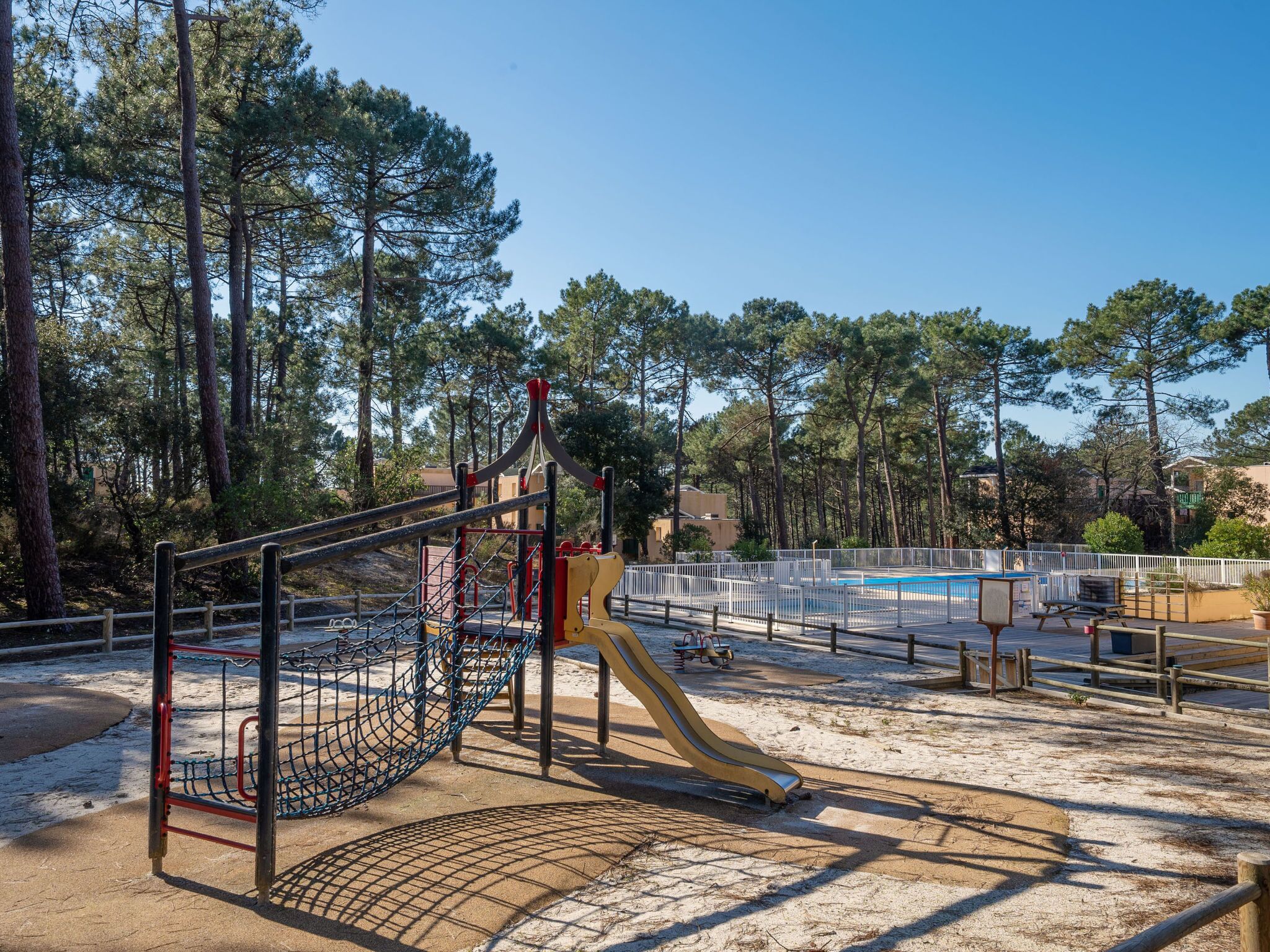 A playground with a slide, climbing net, and swings, surrounded by trees and a pool.