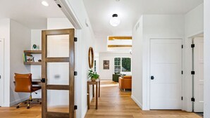 A modern interior with a wooden floor, a desk with a chair, a plant, and a view of a living area with a couch and a window. - Luxury Meets Location | Near Ephraim & Sister Bay (Ephraim)