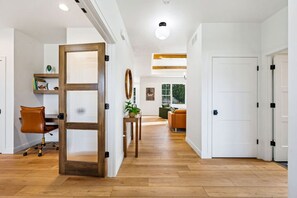 Ein modernes Interieur mit Holzboden, einem Schreibtisch mit Stuhl, einer Pflanze und Blick auf einen Wohnbereich mit Sofa und Fenster.