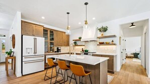 A modern kitchen with wooden cabinets, a white refrigerator, a large island with bar stools, pendant lights, and a view into the adjacent living area. - Luxury Meets Location | Near Ephraim & Sister Bay (Ephraim)