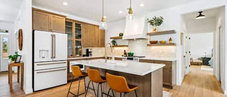A modern kitchen with wooden cabinets, a white refrigerator, a large island with bar stools, pendant lights, and a view into the adjacent living area.