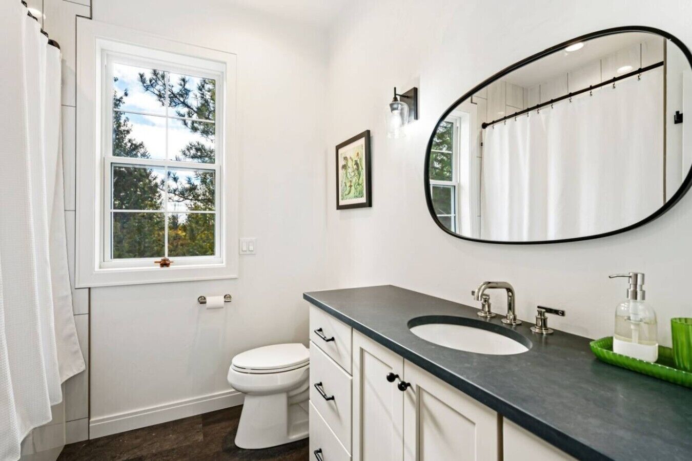 A bathroom with a white toilet, a large mirror, a sink with a dark countertop, and a window with a view of trees.