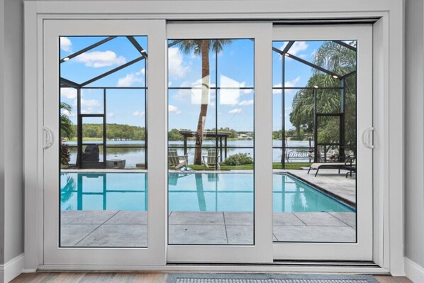 A sliding glass door leading to a swimming pool with a view of a lake and palm trees. - Spacious Lake Tarpon Getaway (Palm Harbor)