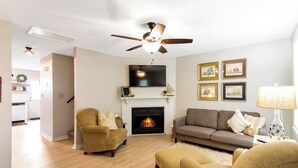 Una sala de estar con chimenea, ventilador de techo, televisor y cuadros en la pared.