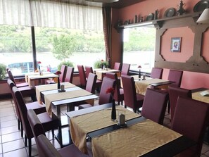 Um restaurante com várias mesas e cadeiras, vista para o verde lá fora e uma parede com pratos decorativos.