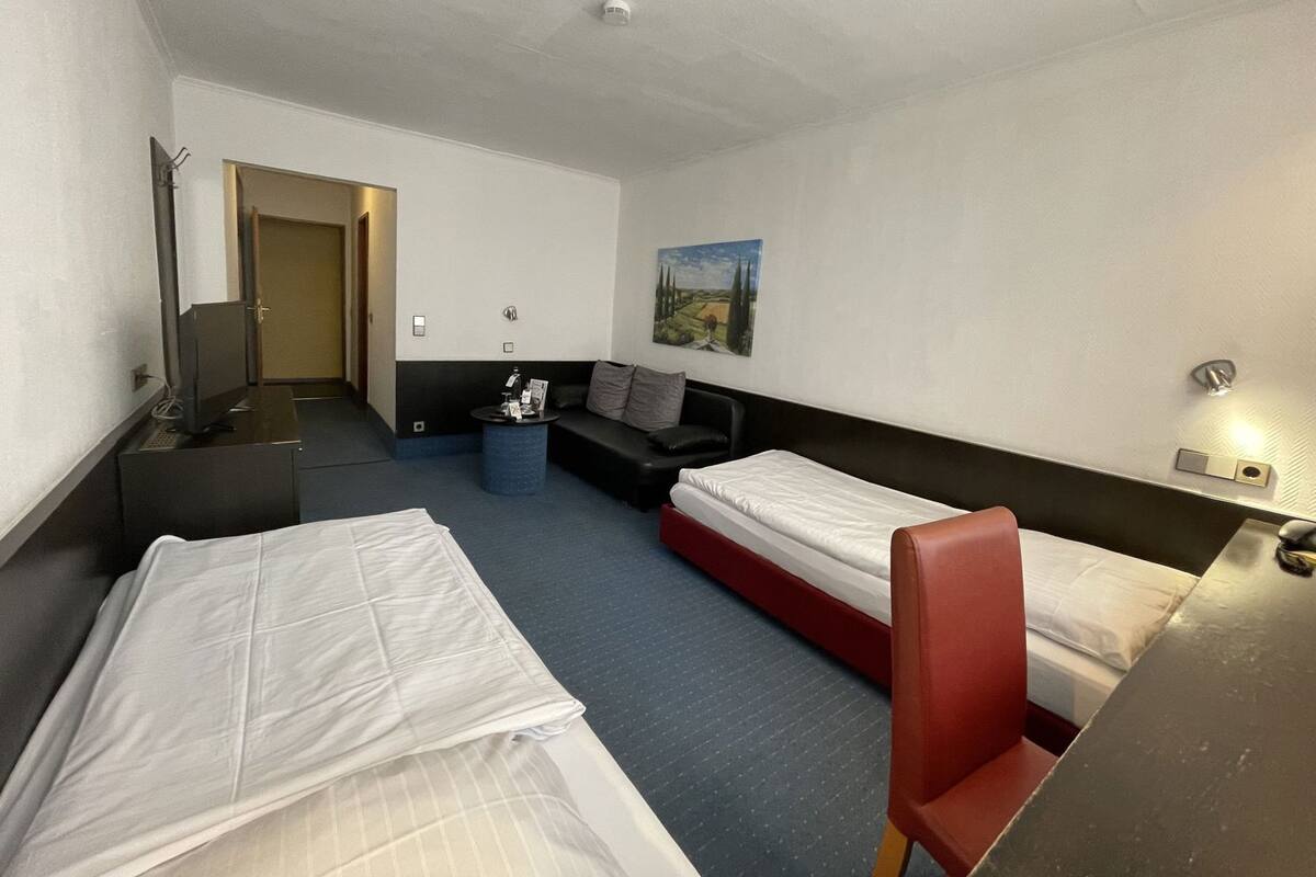 A hotel room with two beds, a desk, a chair, a small table, a sofa, and a painting on the wall.
