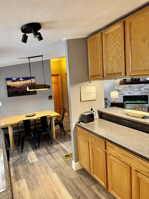 Una cocina moderna con armarios de madera, encimera de granito, lámpara colgante y comedor con mesa y sillas de madera.