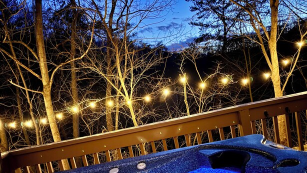 A hot tub with a blue textured surface and multiple control knobs, surrounded by a wooden deck and string lights against a backdrop of trees.