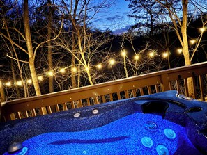 A hot tub with a blue textured surface and multiple control knobs, surrounded by a wooden deck and string lights against a backdrop of trees. - Vista Bear Mineral Cabin w Hot Tub, Game Room & WiFi close to attractions  (Sevierville)