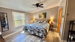 A bedroom with a bed, a ceiling fan, a window with blinds, a painting on the wall, and a nightstand with a lamp.