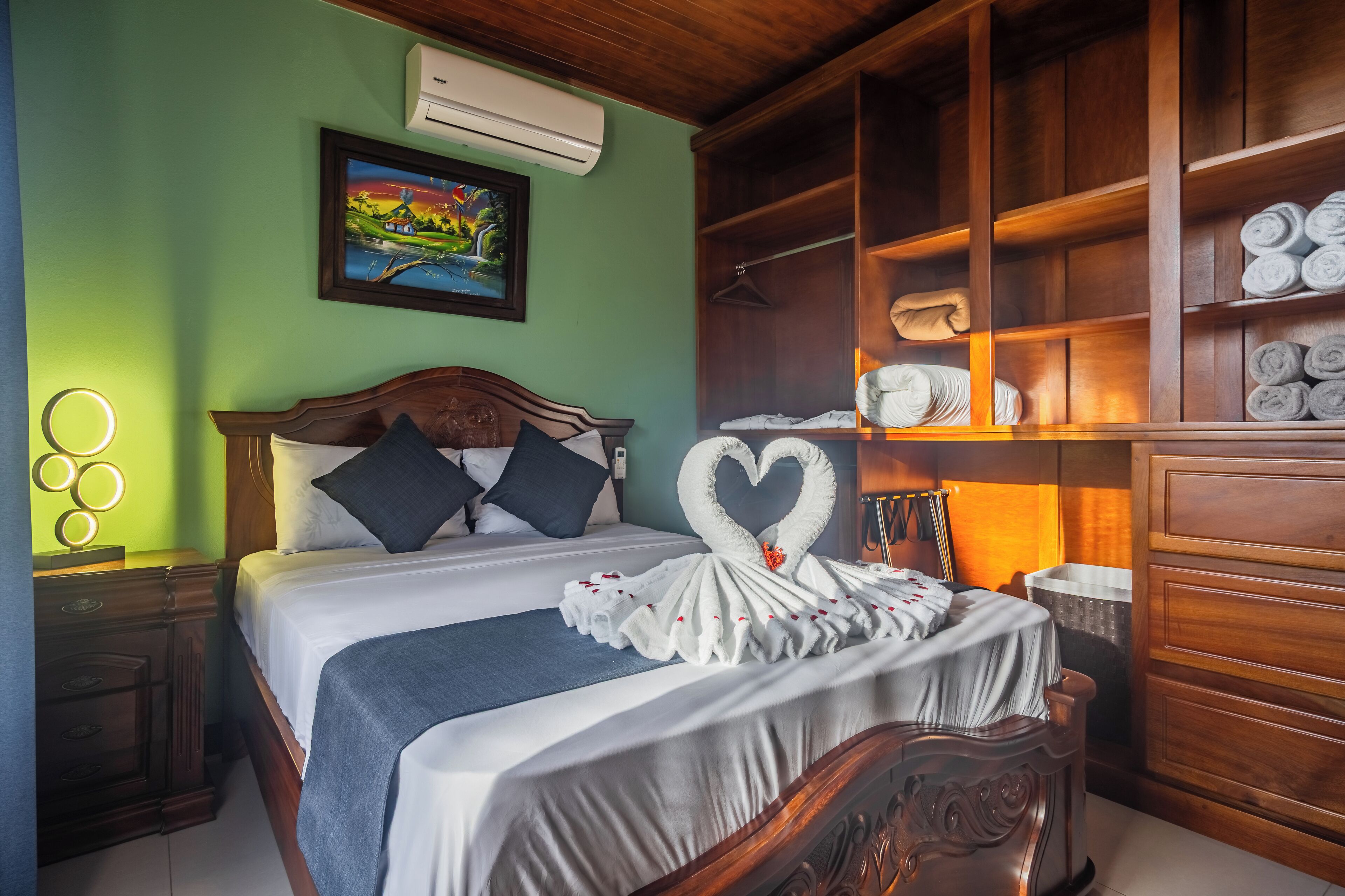 A hotel room with a bed, a wooden headboard, a wall-mounted air conditioner, a painting, a wardrobe with towels, and a bedside table with a lamp.