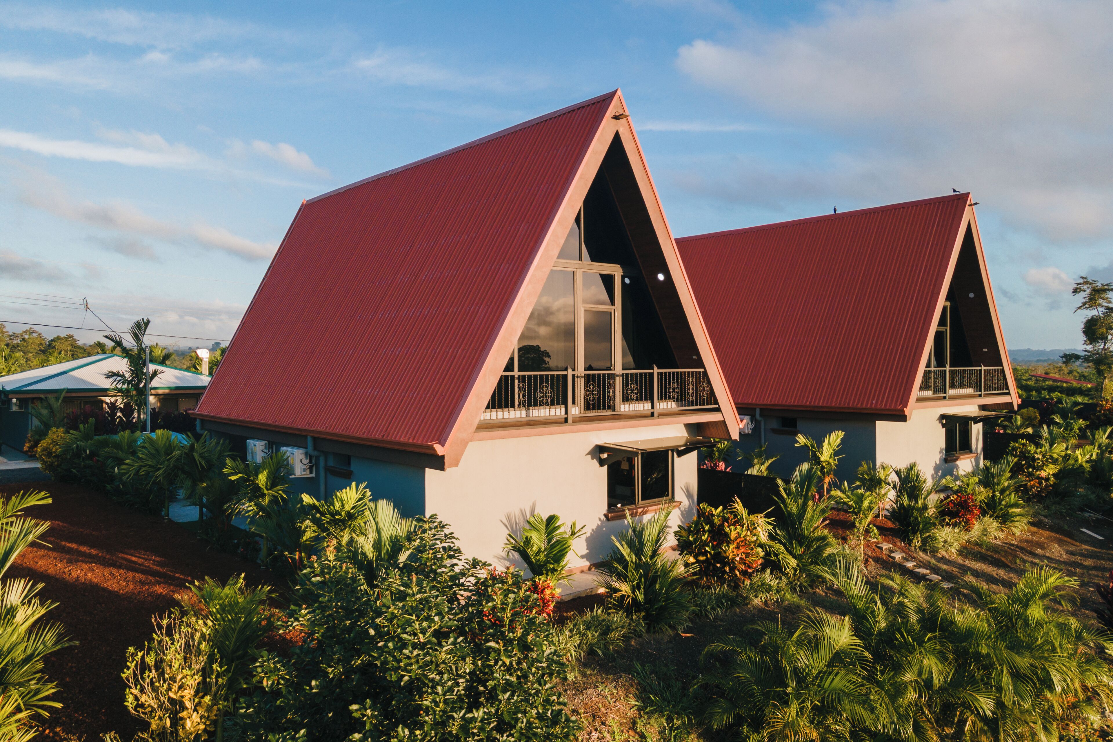 A modern house with a red roof and large windows, surrounded by lush greenery.