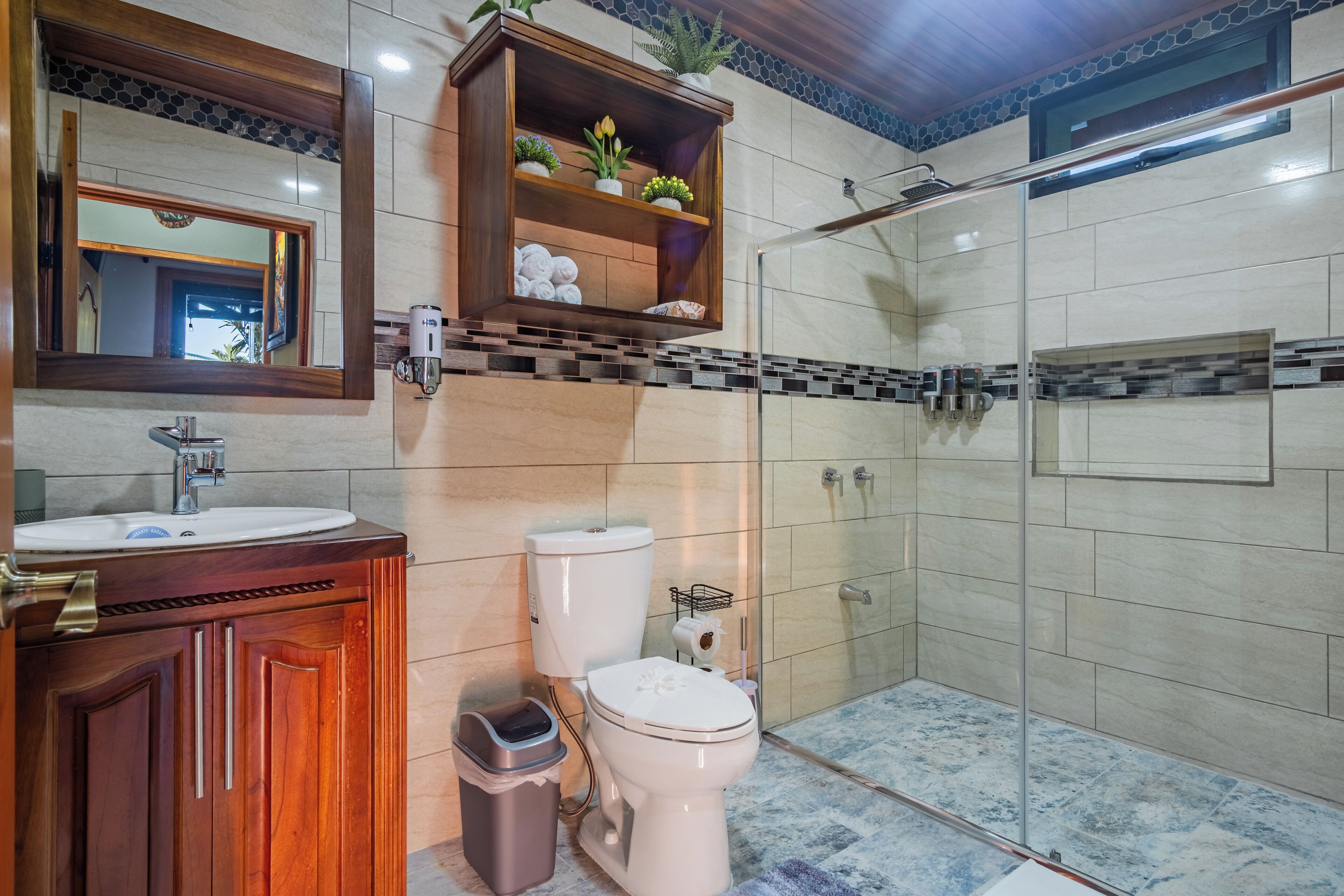 Une salle de bain avec un meuble-lavabo en bois, des toilettes, une douche en verre et un miroir avec une étagère au-dessus.