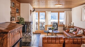 A cozy living room with a wooden ceiling, a stone fireplace, a leather sofa, and a view of a snowy landscape through large windows.