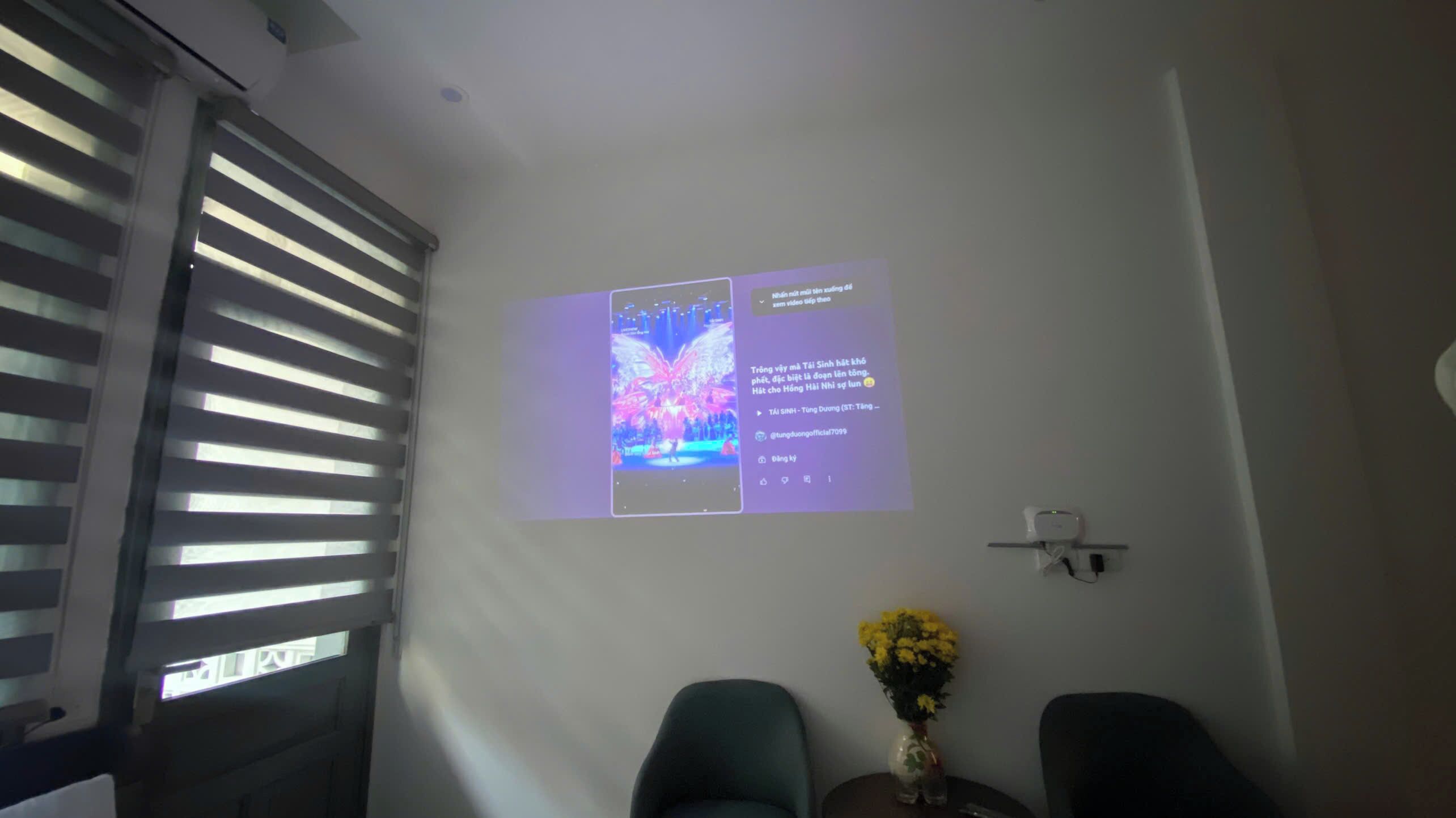 Une pièce équipée d’un écran de projection diffusant une vidéo, d’une fenêtre avec des stores et d’une petite table sur laquelle est posé un vase de fleurs.