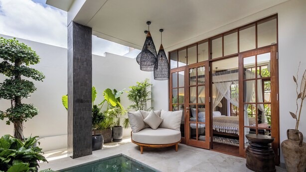 A modern outdoor pool area with a sofa, potted plants, and a wooden door leading to a bedroom. - Plawa Laguna Bali (Pererenan)