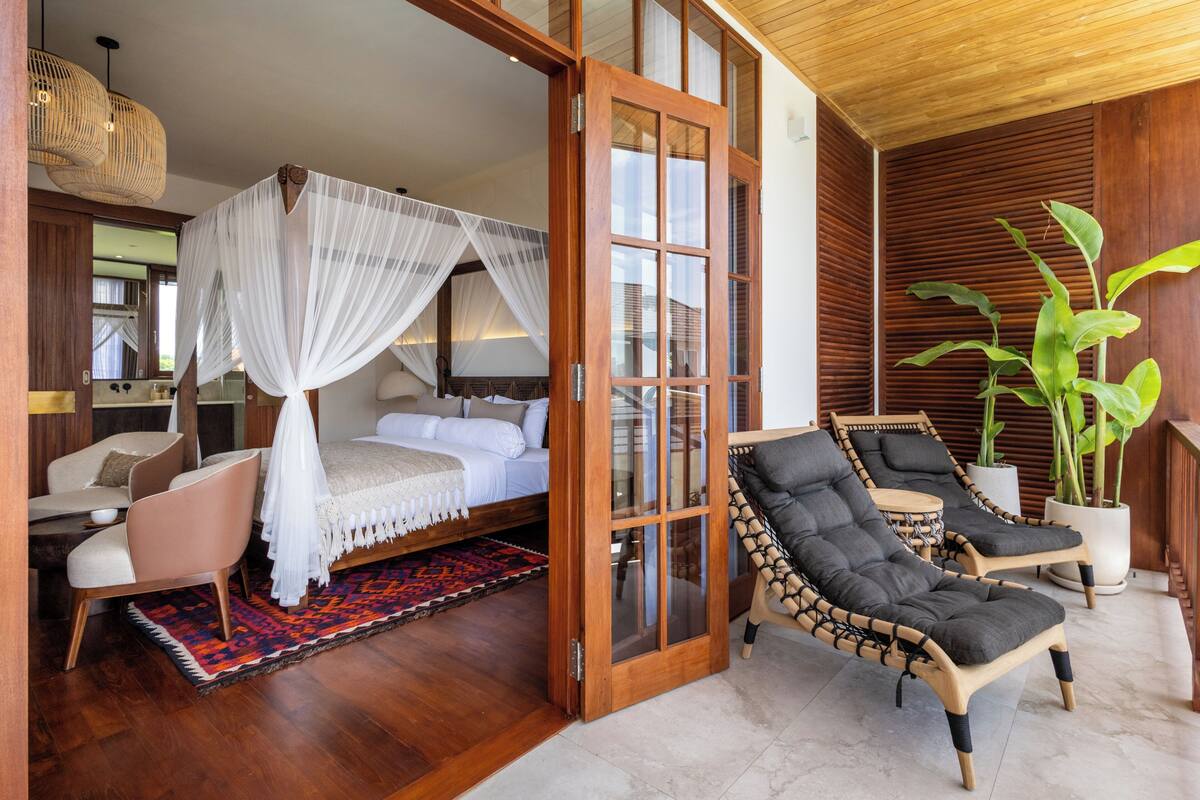 A bedroom with a bed, a sitting area with chairs, a large window, and a potted plant.