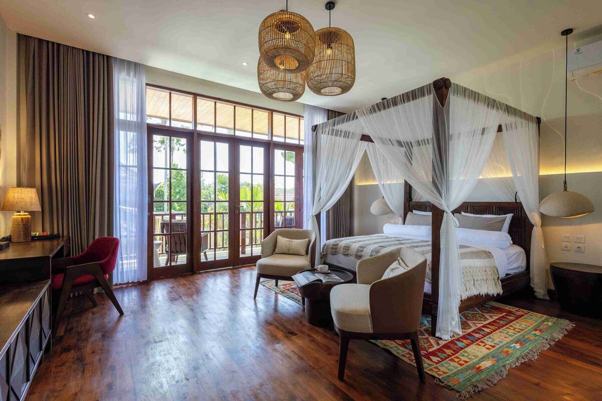 A spacious bedroom with a four-poster bed, a sitting area with chairs, a large window with curtains, and a wooden floor.