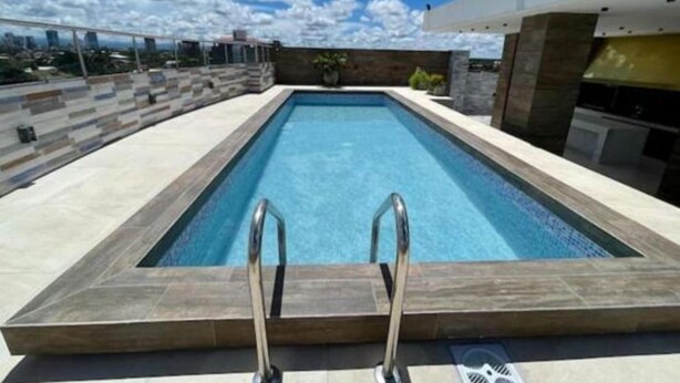 Une piscine rectangulaire avec des échelles métalliques à la partie peu profonde, entourée d’un mur de pierre et avec un bâtiment moderne en arrière-plan.